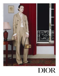 Model wearing a beige trench coat and matching trousers, holding a leopard print handbag in a room with red curtains.