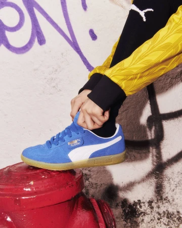 Blue Puma Palermo sneaker with white stripe sits atop a red fire hydrant with graffiti in the background, street-style footwear.