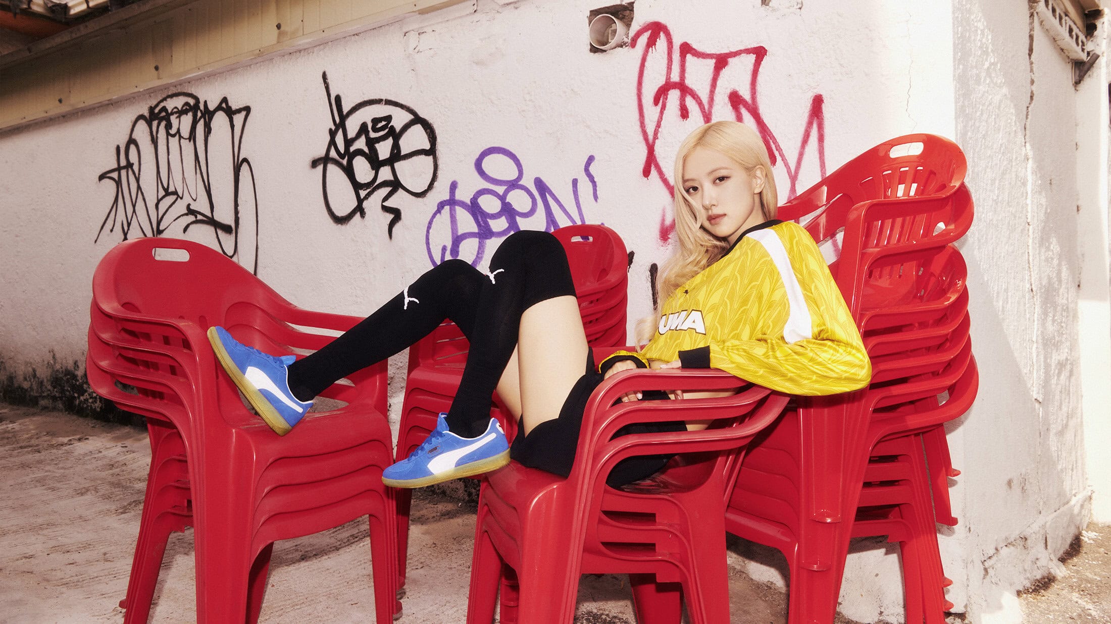 Blonde model in yellow jacket poses on stack of red chairs with graffiti wall background, wearing Puma shoes and black knee-high socks.