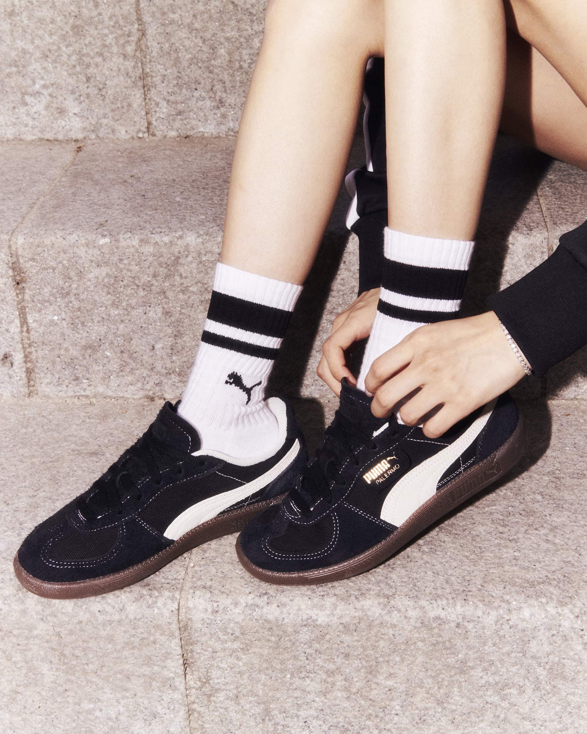 Close-up of woman sitting on steps, wearing black and white Puma sneakers with striped socks, tying the laces on one of the shoes.