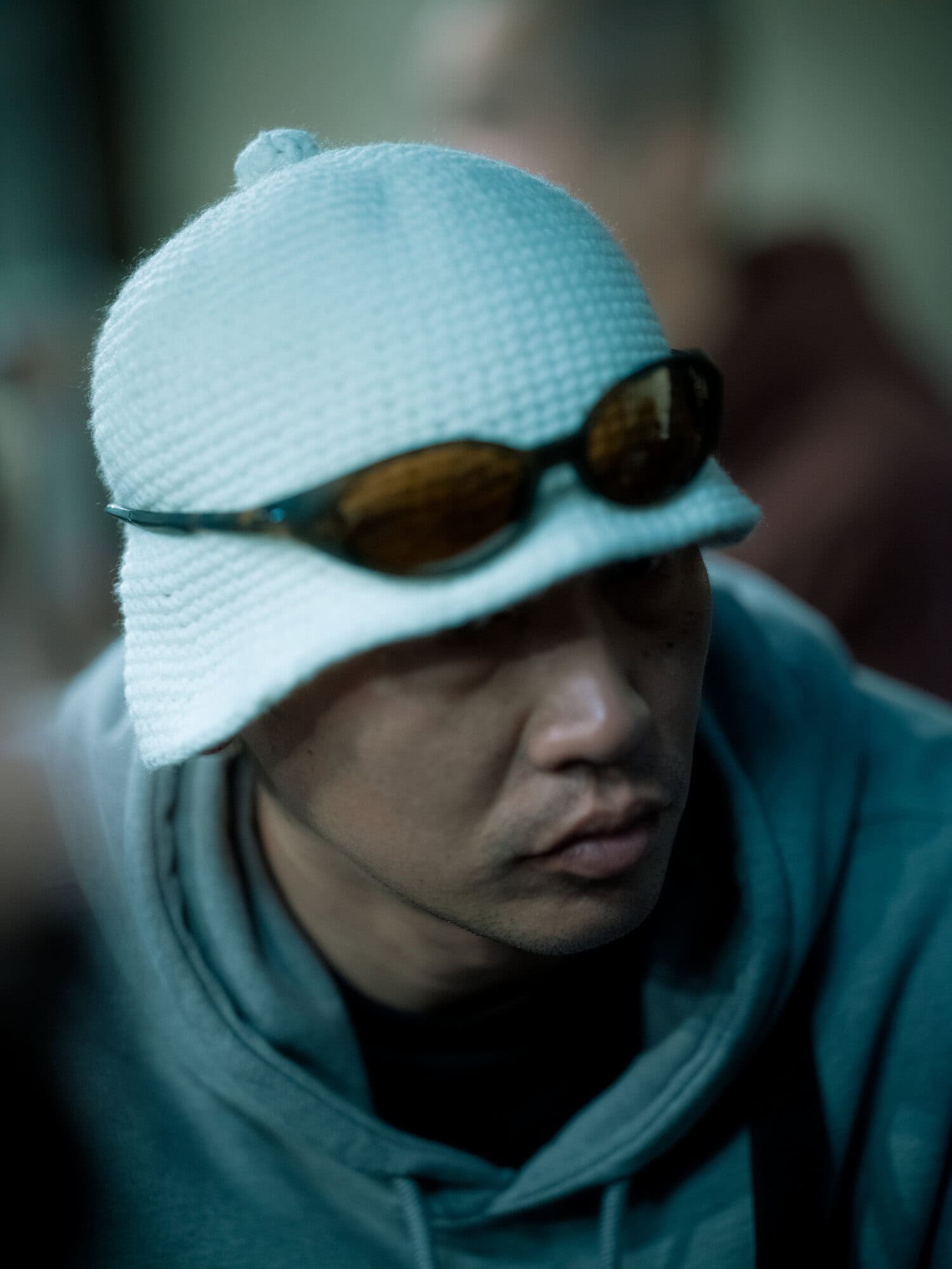 Close-up of a person wearing a white knit hat and sunglasses, eyes are partially obscured, wearing a grey hooded sweatshirt, indoors.
