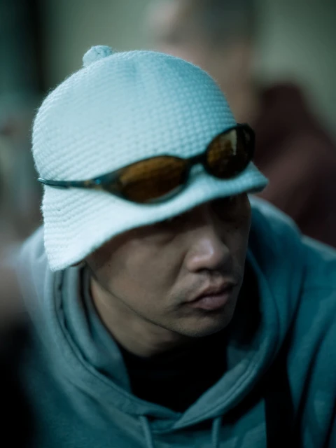 Close-up of a person wearing a white knit hat and sunglasses, eyes are partially obscured, wearing a grey hooded sweatshirt, indoors.