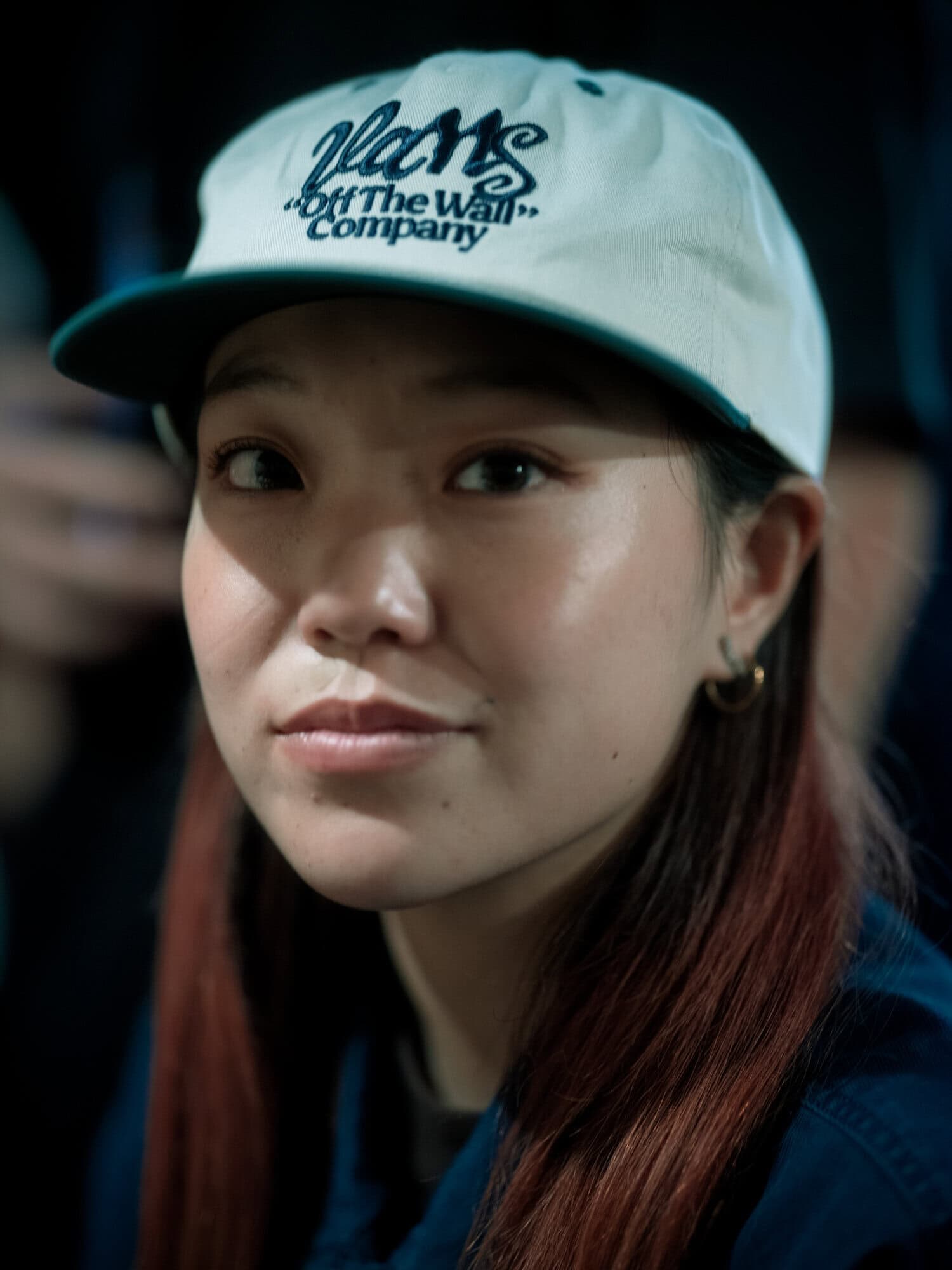 Close-up of a smiling Asian woman in a white Vans cap, shot indoors with soft lighting. The focus is on her face and expression.