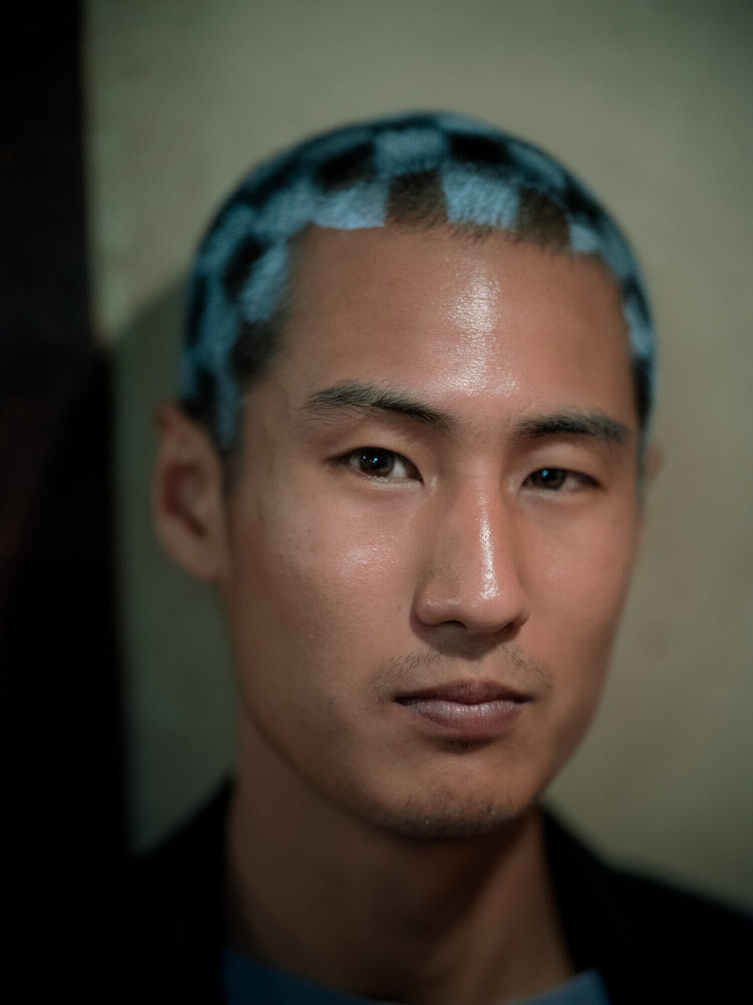 Close-up portrait of an adult man with short blue and black checkered hair, gazing forward with a neutral expression in a dimly lit indoor space.