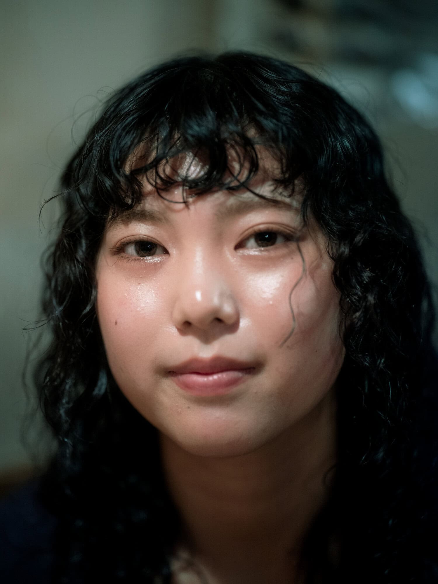 Close-up portrait of a young adult woman with curly dark hair, fair skin, and a neutral expression, softly lit indoors.
