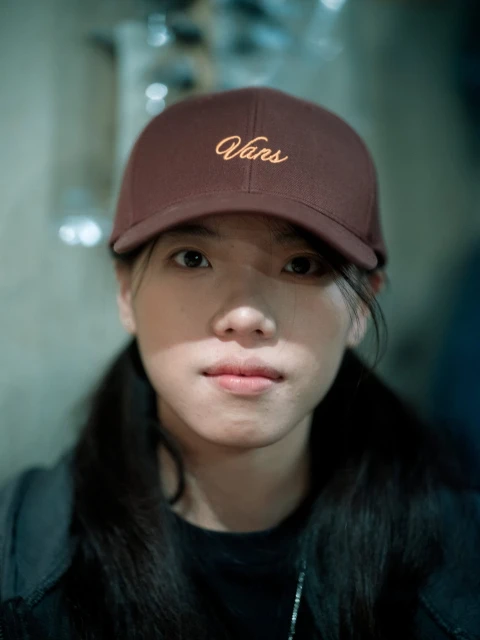 Portrait of a young Asian woman wearing a brown Vans baseball cap, looking directly at the camera in an indoor environment.