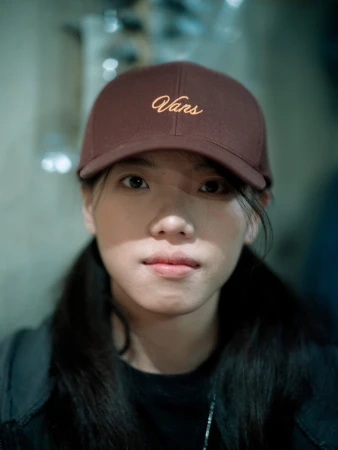 Portrait of a young Asian woman wearing a brown Vans baseball cap, looking directly at the camera in an indoor environment.