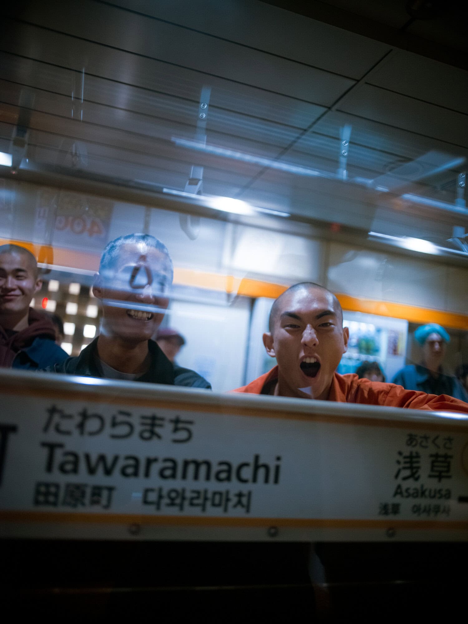 Three bald men in a subway station, one yelling, another smiling, and a third partially visible, all reflected in a station sign.