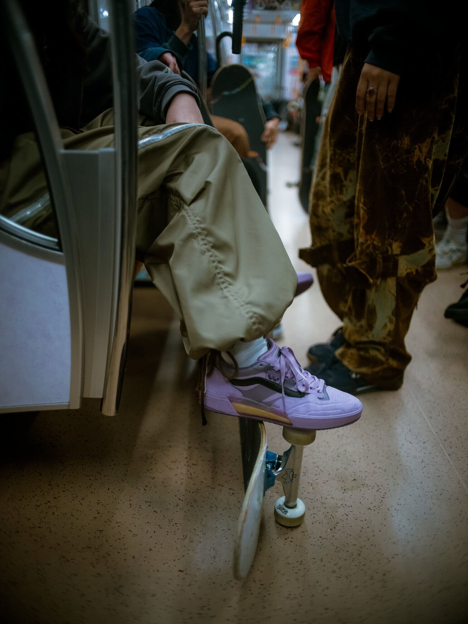 Person riding public transport with a skateboard and light purple sneakers, wearing beige pants, with other passengers nearby.