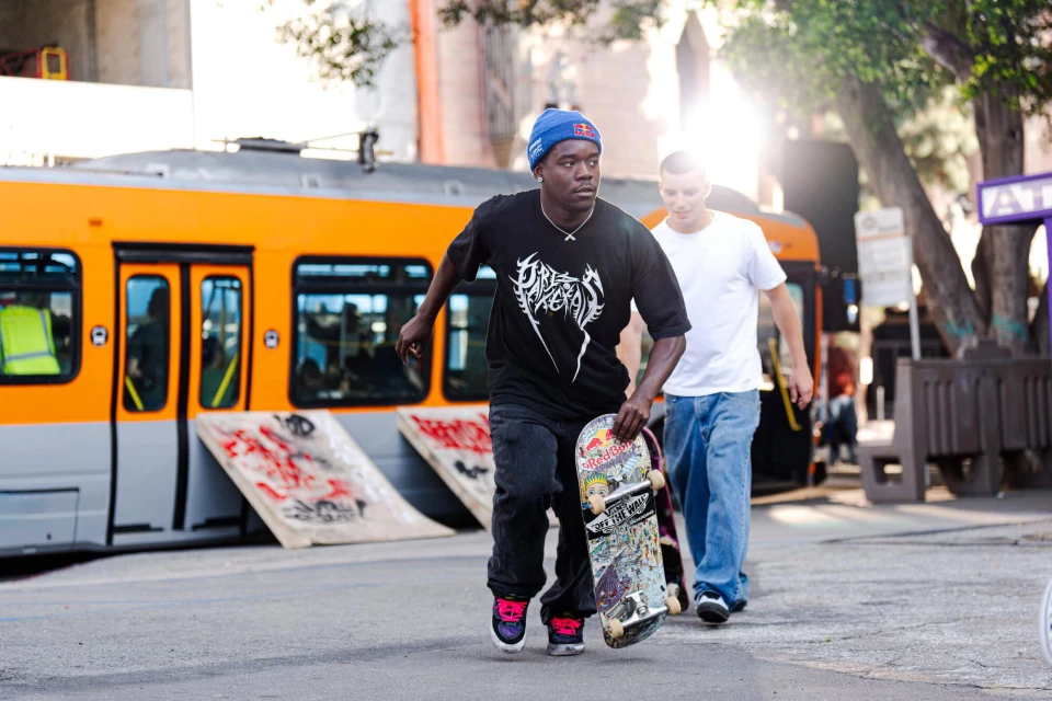 A skateboarder with a blue beanie walks toward the camera, holding his skateboard. Another man and an orange train car are behind him.