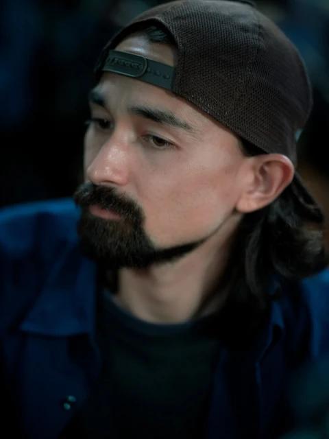 Close-up portrait of an adult male with a goatee and long hair wearing a baseball cap. Focused expression and casual blue jacket.