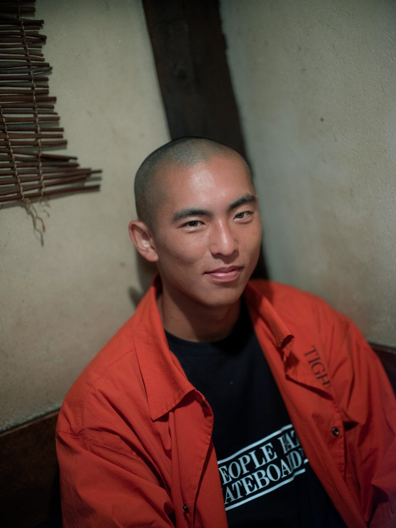 Portrait of an Asian man with a shaved head indoors, wearing an orange collared shirt over a black t-shirt with white text.