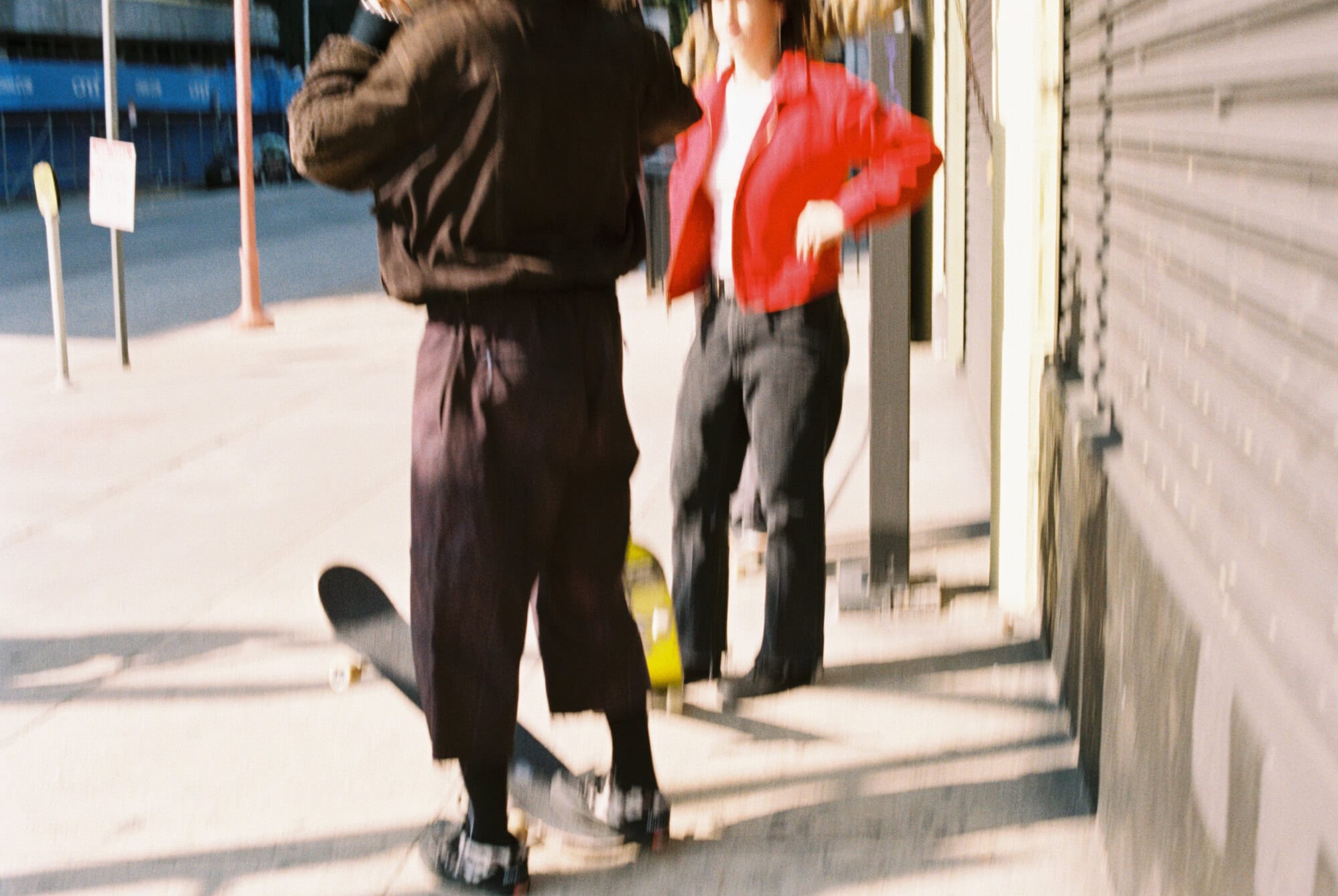 Two people stand on a sidewalk next to a skateboard in an urban setting with a red jacket, dark pants, and a brown jacket. 