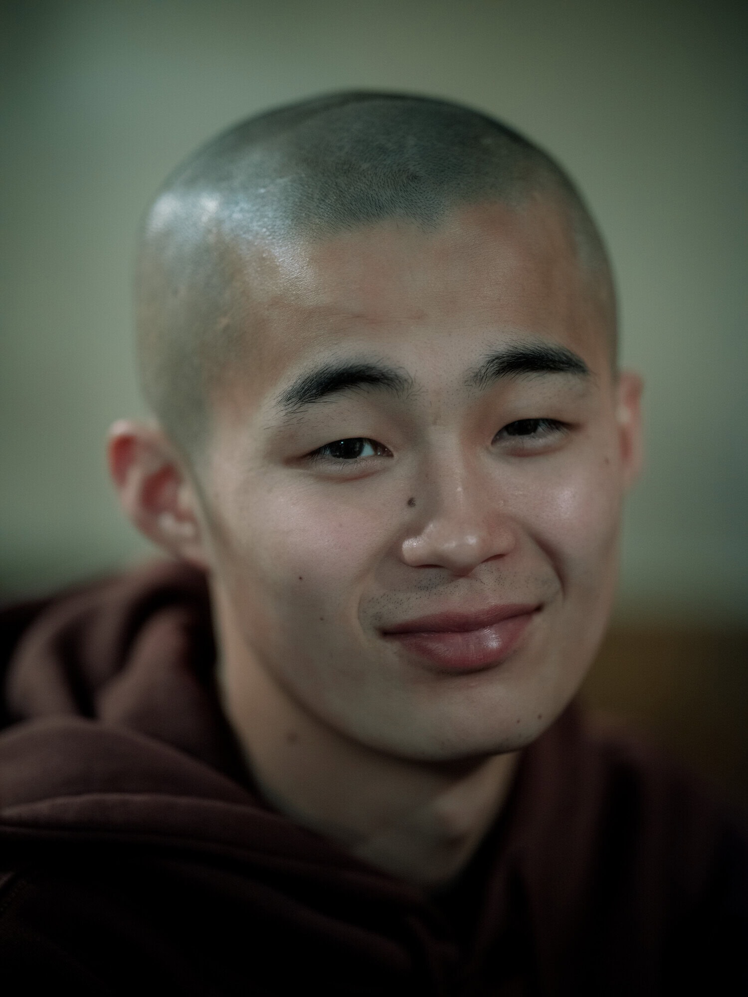 Close-up portrait of a young adult with a shaved head, fair skin, and a gentle smile, wearing a brown hooded sweatshirt indoors.