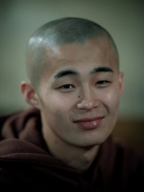 Close-up portrait of a young adult with a shaved head, fair skin, and a gentle smile, wearing a brown hooded sweatshirt indoors.