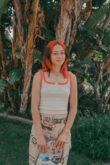 Portrait of a young woman with dyed hair, a tank top, and a Vans skateboard, posing in front of a lush green tropical plant.