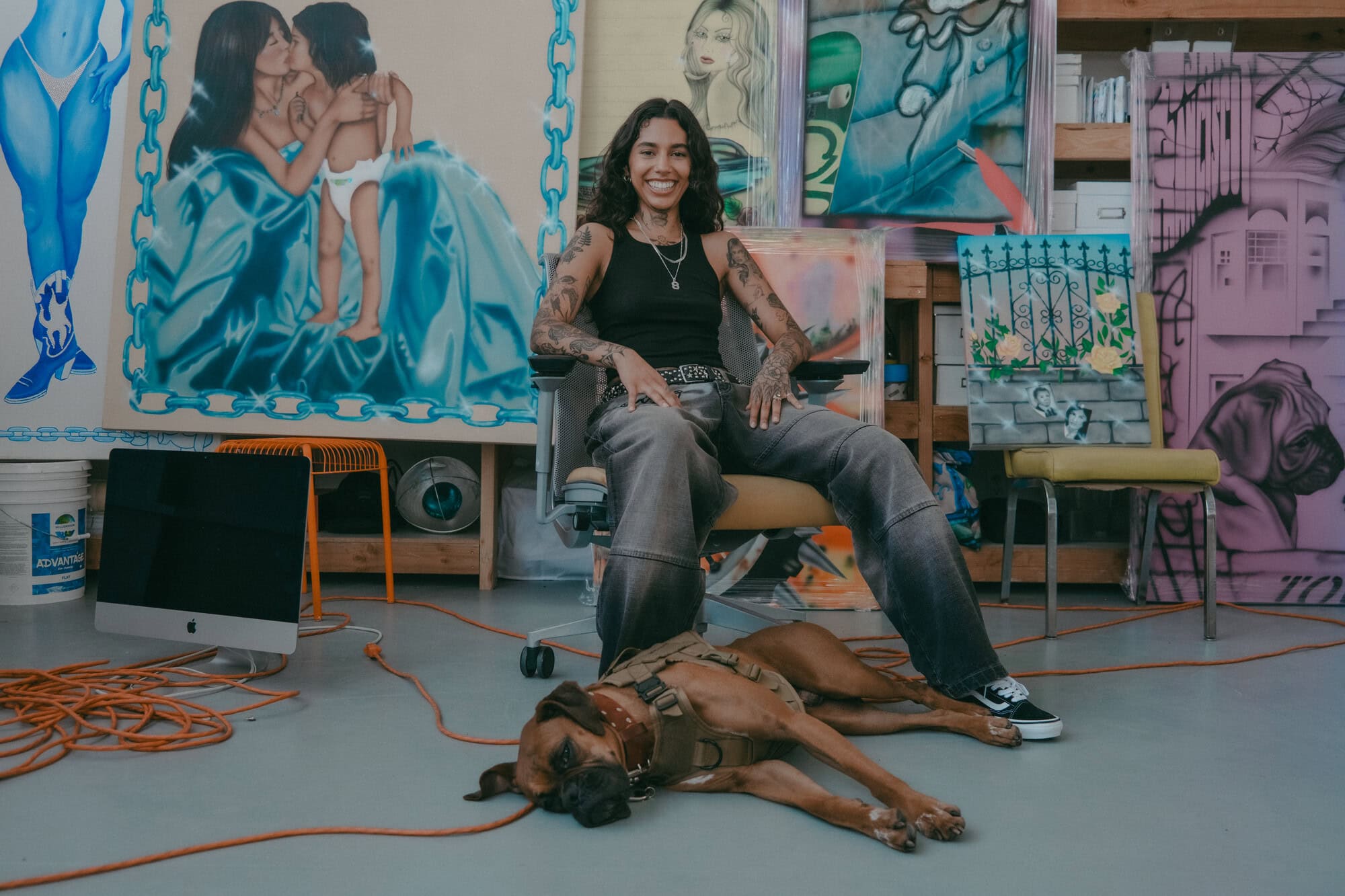 Tattooed artist sits with her dog in her colorful studio amid her paintings, surrounded by an iMac computer and art supplies.