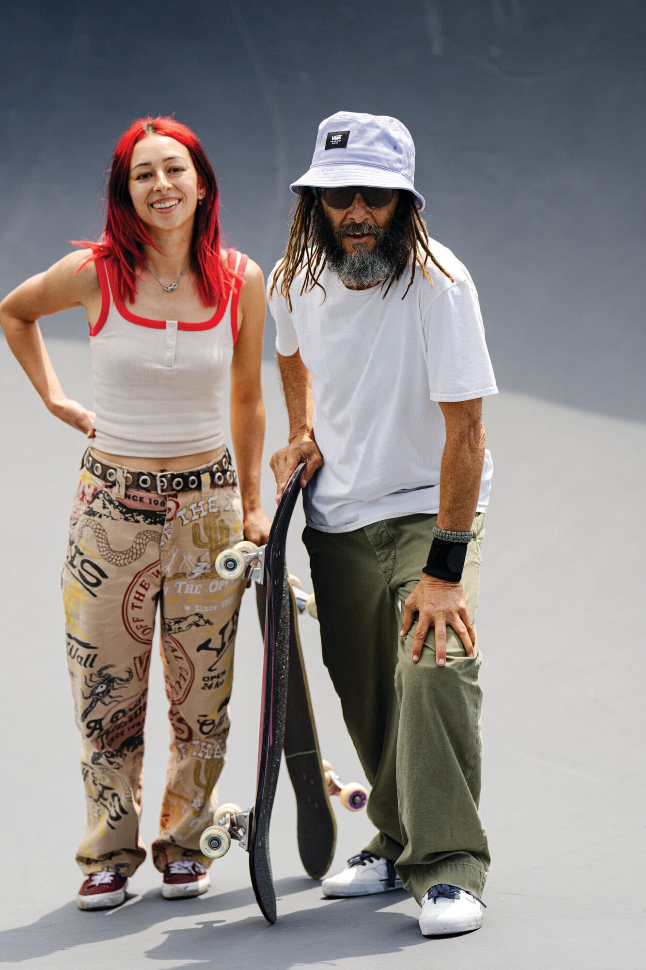 Two skateboarders, an adult female and adult male, stand at a skatepark. The female has long red hair; the male wears a bucket hat.