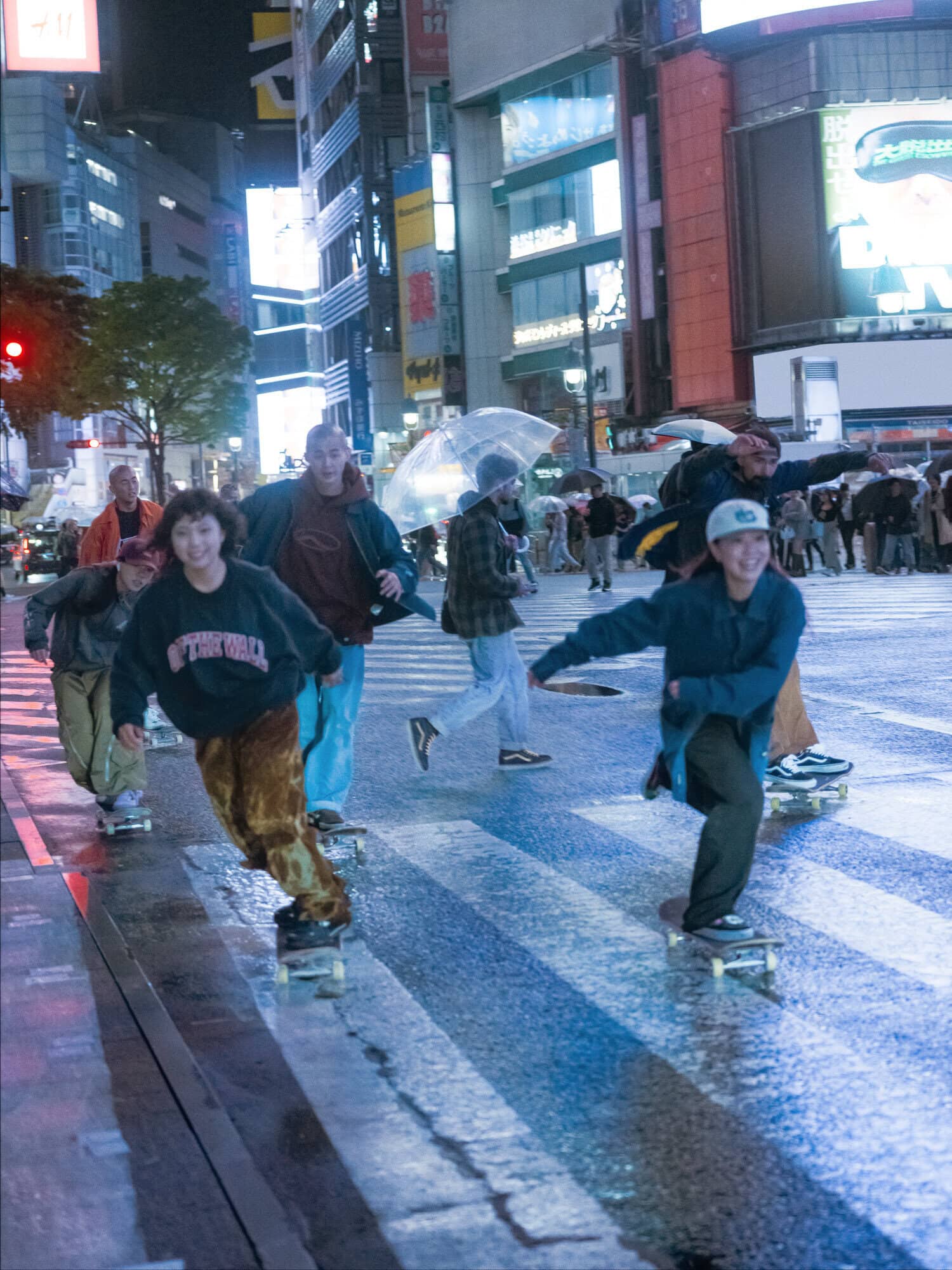 Group of young adults skateboarding on a wet city street at night, illuminated by bright buildings and traffic lights in an urban setting.