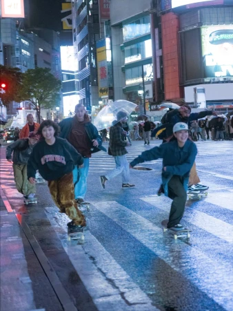 Group of young adults skateboarding on a wet city street at night, illuminated by bright buildings and traffic lights in an urban setting.