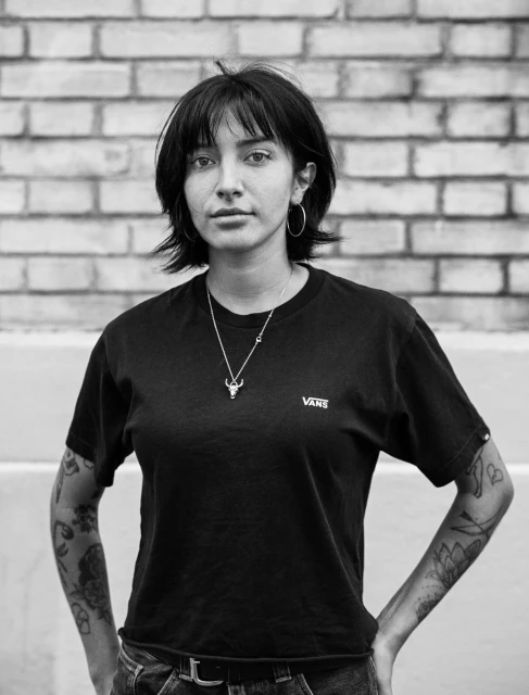 Black and white portrait of a young woman with dark hair and tattoos, wearing a black t-shirt and standing in front of a brick wall.