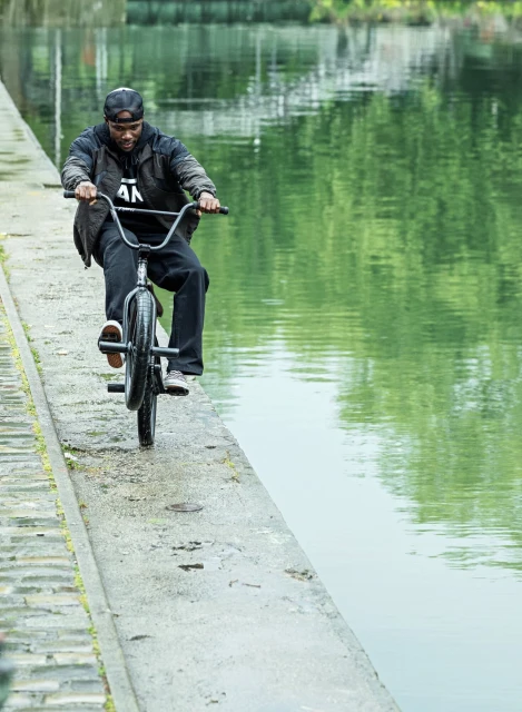 A man riding a BMX bike on a narrow concrete path next to a canal, wearing a hat, jacket, and pants; water and green foliage are visible.