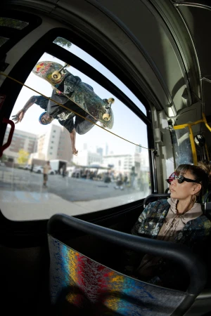 Low-angle shot inside bus with a skateboarder performing aerial trick seen through the window, and a woman passenger watching.