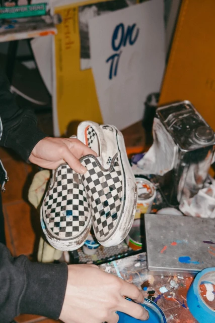 Close-up of a person holding a pair of checkerboard Vans slip-on sneakers over a cluttered table in an indoor setting.