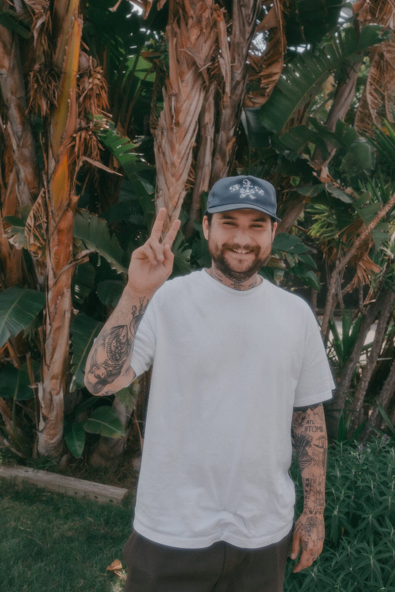 Smiling adult male with tattoos posing outdoors with a peace sign, wearing a white t-shirt, cap, and surrounded by lush green foliage.