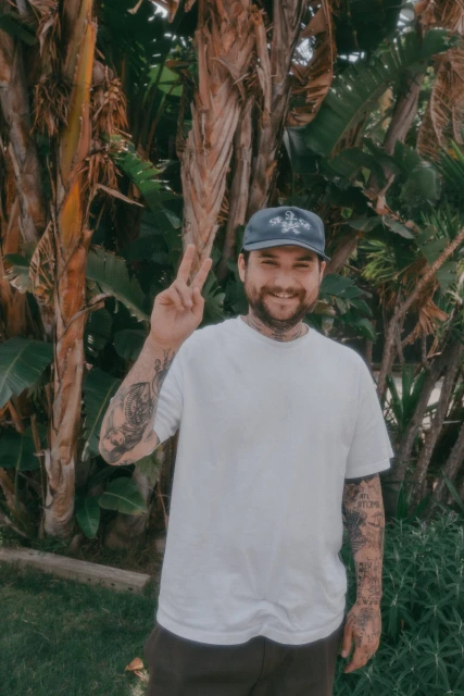 Smiling adult male with tattoos posing outdoors with a peace sign, wearing a white t-shirt, cap, and surrounded by lush green foliage.