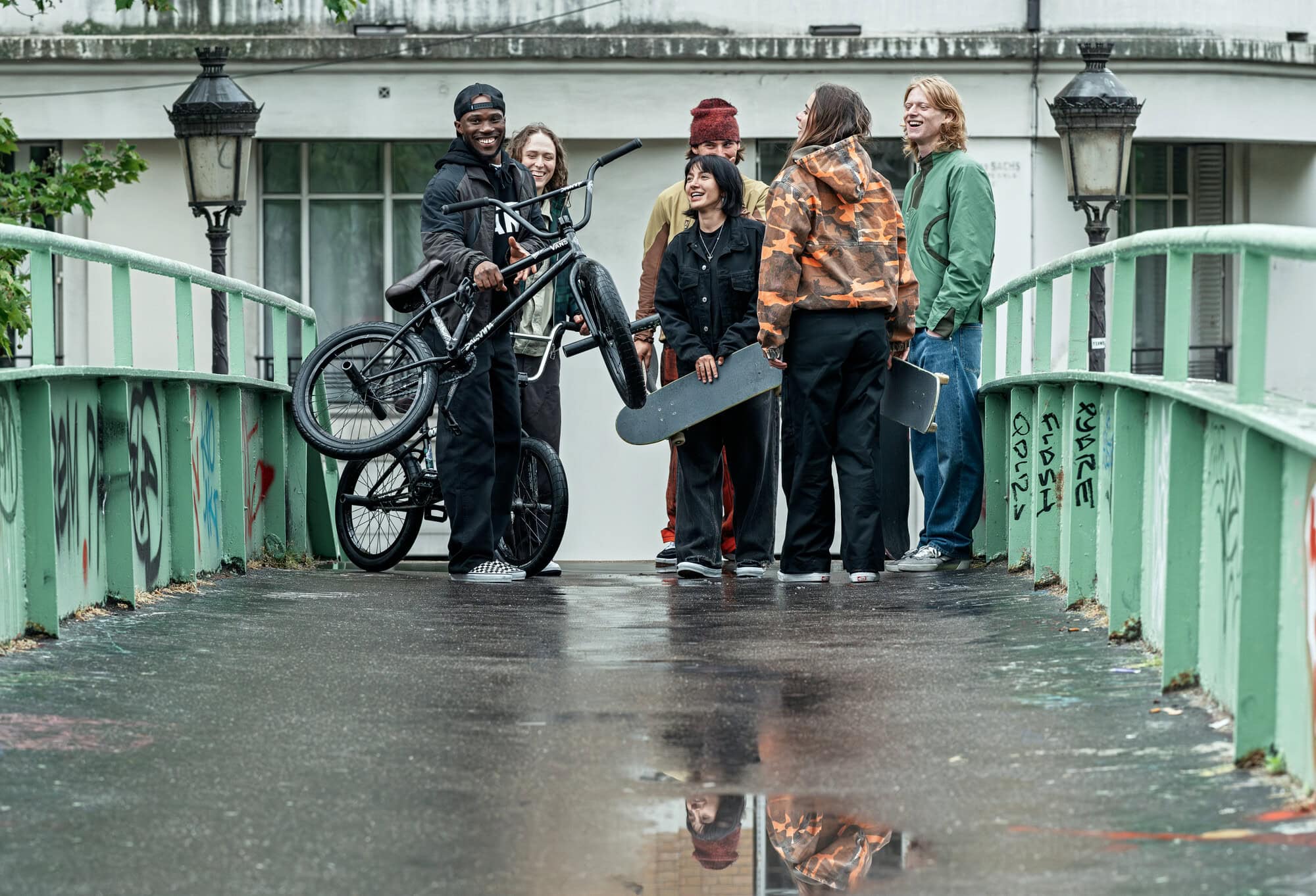 Group of five young adults gathered on a bridge, one carrying a bicycle, another a skateboard, urban setting with muted colors.