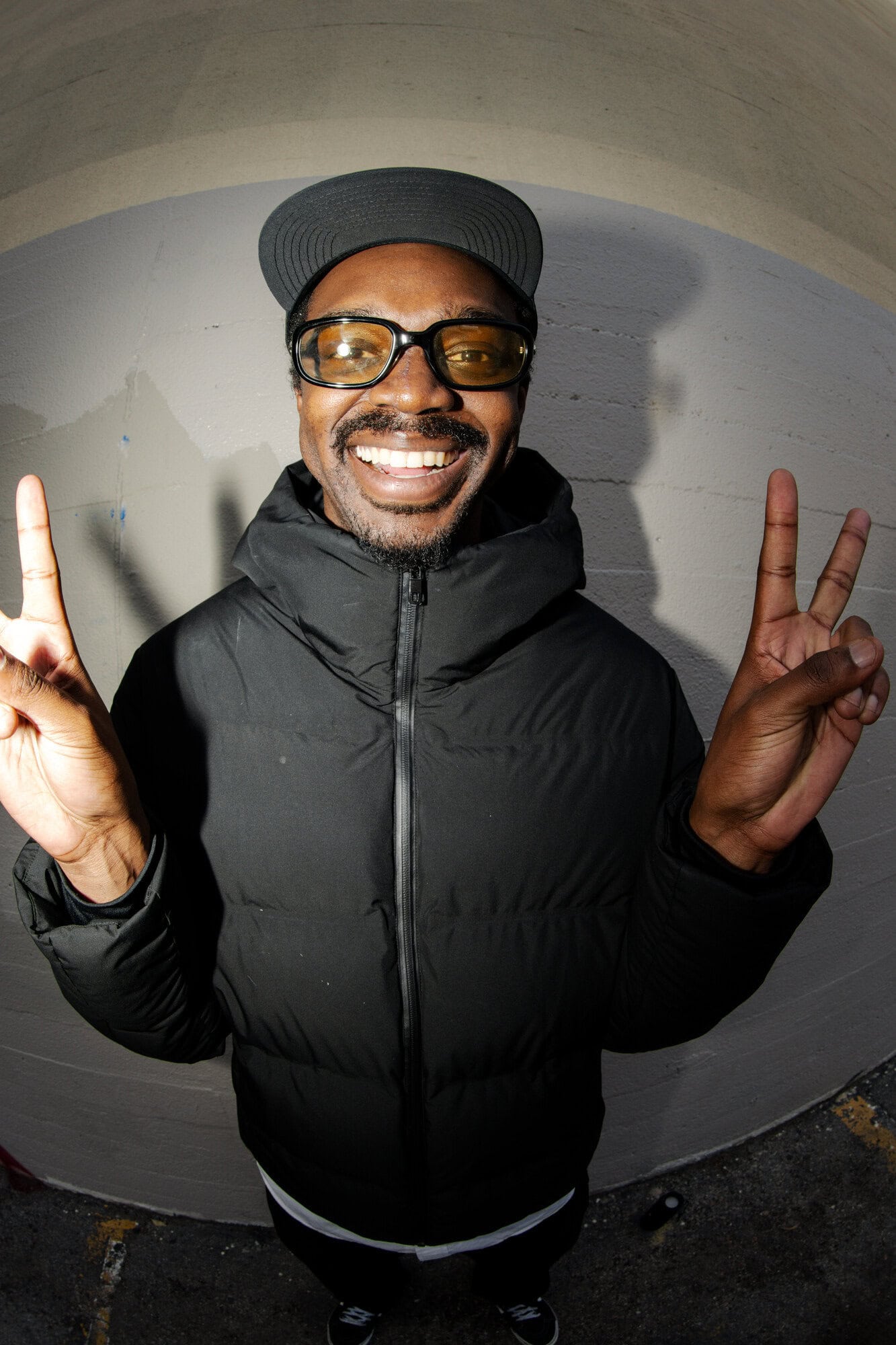 Portrait of a smiling adult male wearing glasses and a black puffer jacket, making peace signs with both hands in an arched tunnel.