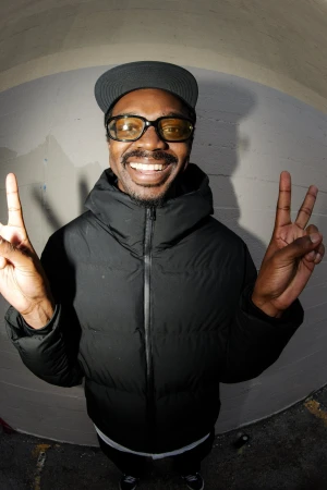 Portrait of a smiling adult male wearing glasses and a black puffer jacket, making peace signs with both hands in an arched tunnel.