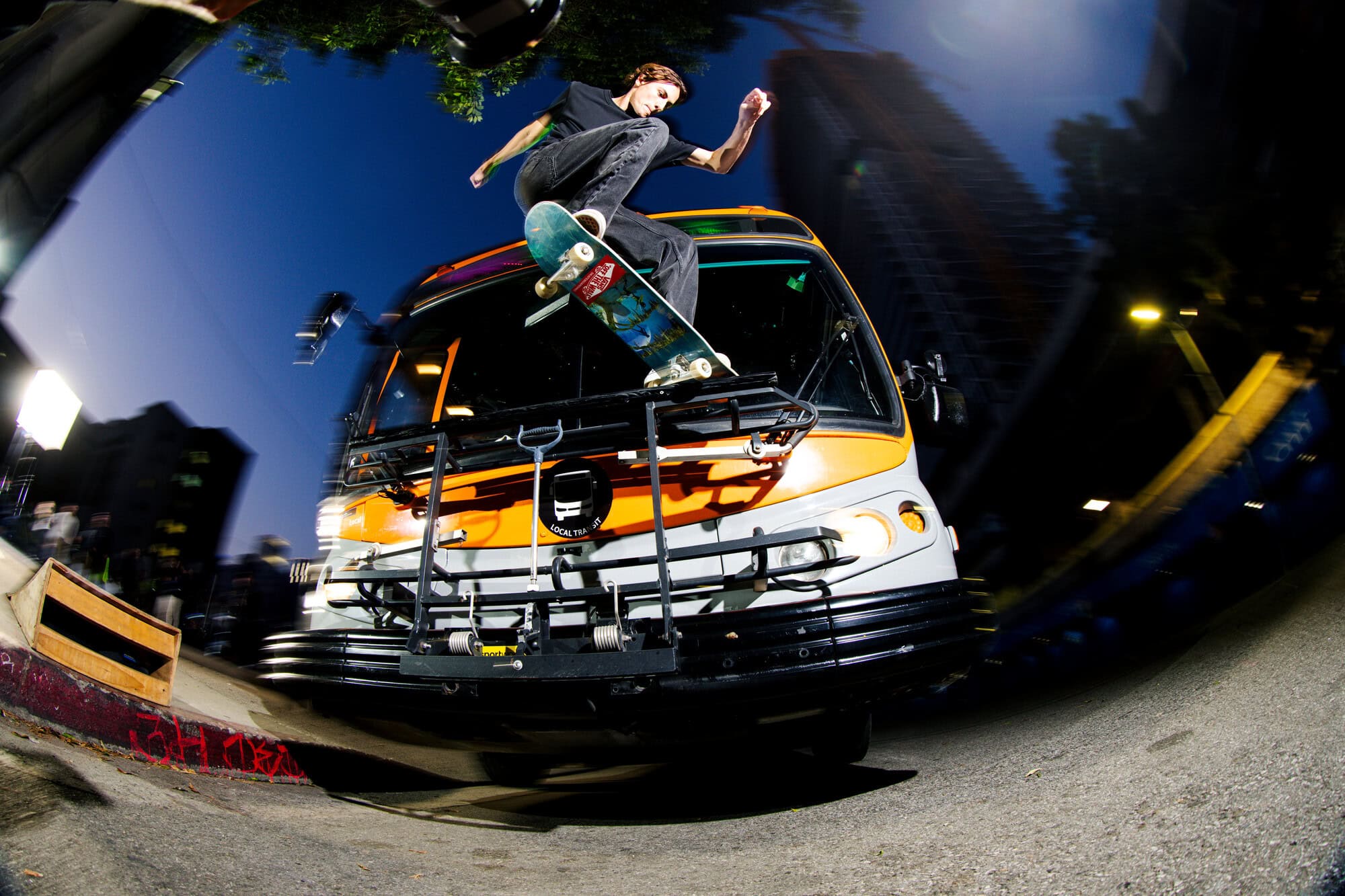 Skateboarder performs a trick on top of a bus in an urban environment with a fisheye lens, capturing the dynamic action and the city backdrop.