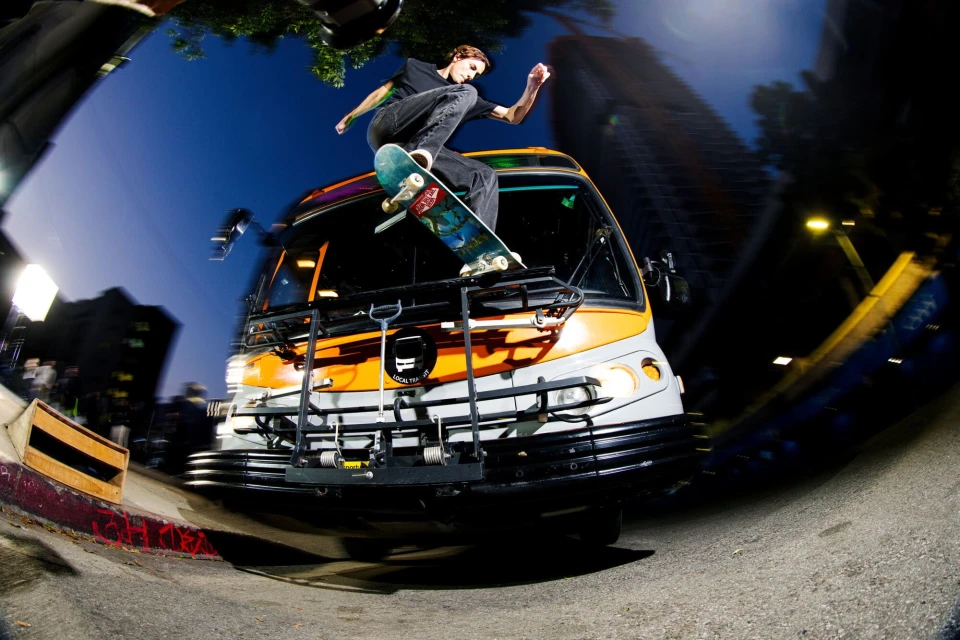 Skateboarder performs a trick on top of a bus in an urban environment with a fisheye lens, capturing the dynamic action and the city backdrop.