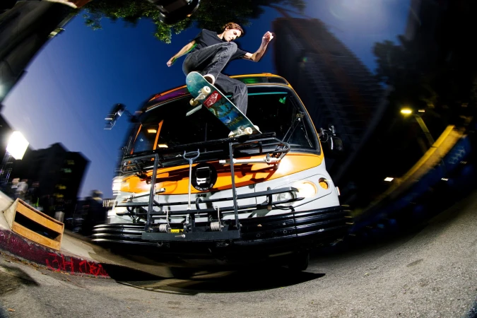 Skateboarder performs a trick on top of a bus in an urban environment with a fisheye lens, capturing the dynamic action and the city backdrop.