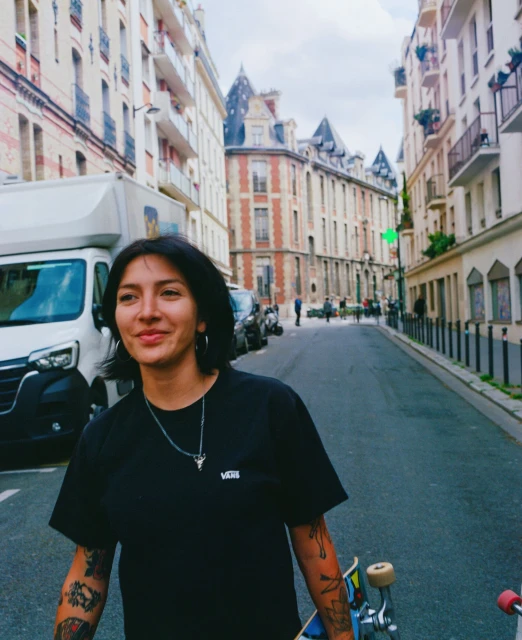 Young woman with tattoos and skateboard walks on street in Paris, France, near classic architecture. Wearing a Vans t-shirt and silver jewelry.