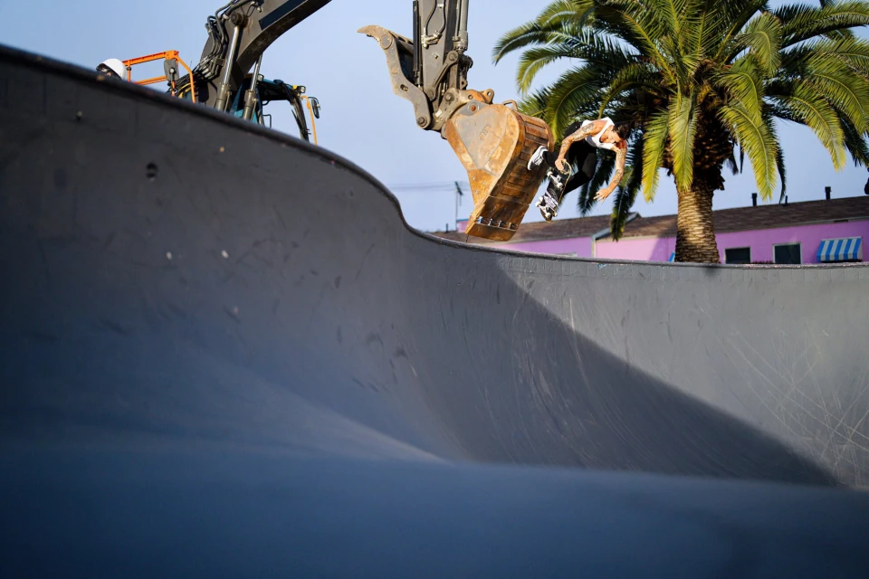 A skateboarder performs a trick in a skatepark bowl, with an excavator bucket suspended above, palm tree and building in the backdrop.