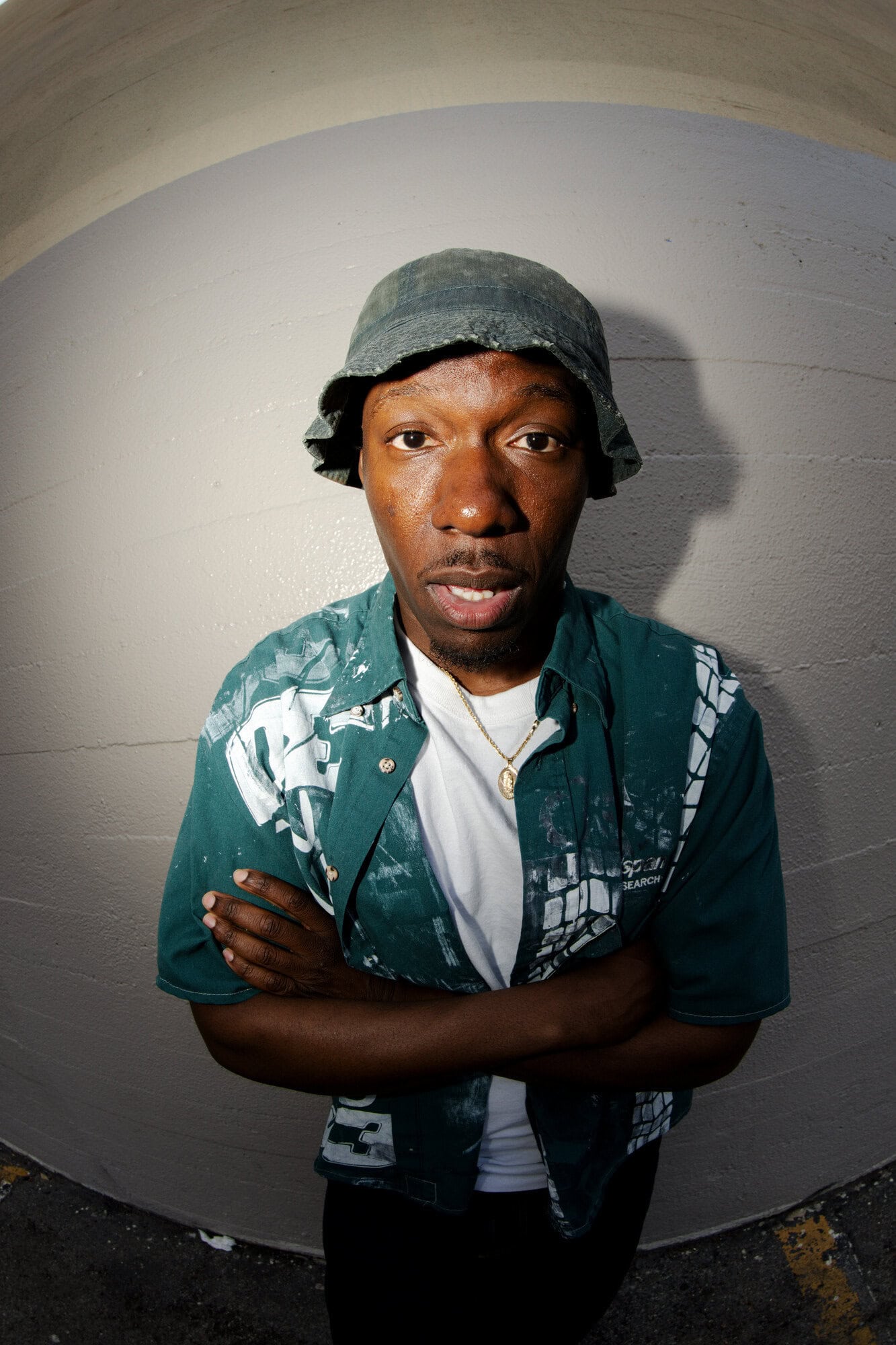 Portrait of a man wearing a bucket hat and patterned shirt standing indoors with arms crossed, captured with a fisheye lens.