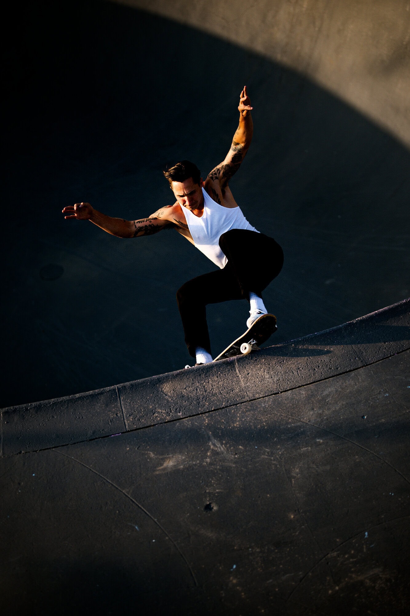 A skateboarder in a white tank and dark pants soars in a concrete skatepark bowl, capturing a dynamic moment mid-air.