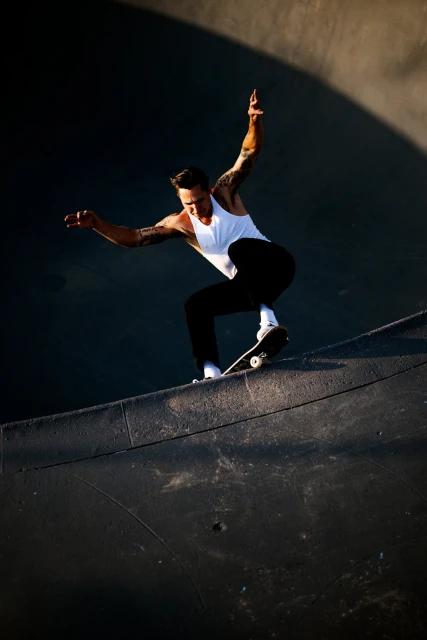 A skateboarder in a white tank and dark pants soars in a concrete skatepark bowl, capturing a dynamic moment mid-air.