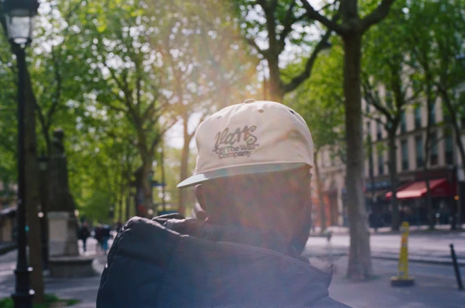 Rear view of a person wearing a white Vans cap and dark jacket on a tree-lined city street with warm sunlight filtering through branches.