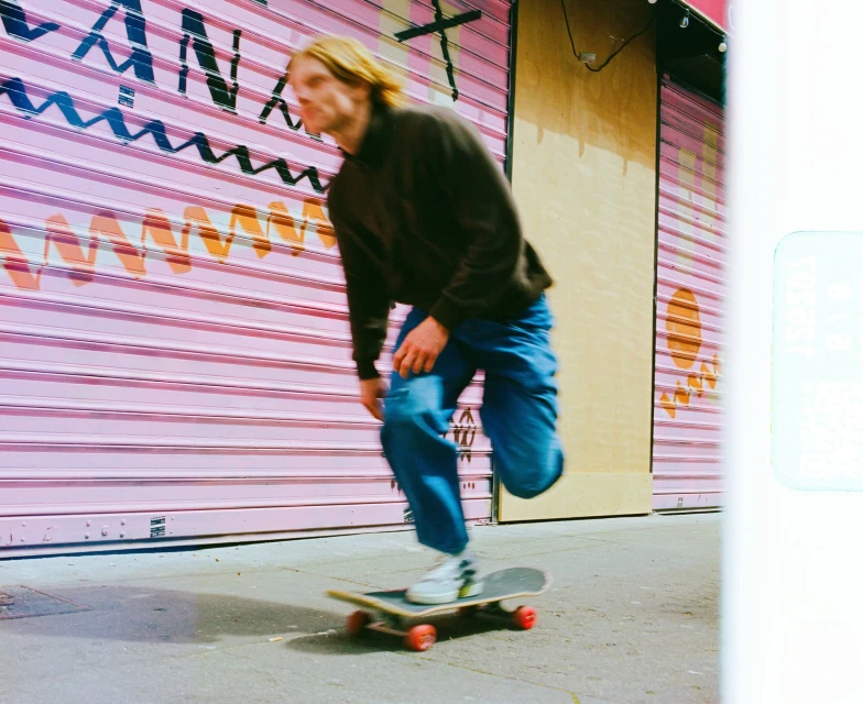 A person rides a skateboard on a city street, past a pink graffitied storefront. The man has long hair and wears jeans and a jacket.