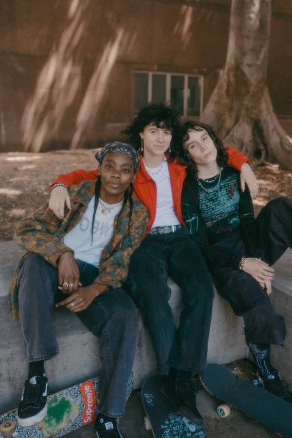 Three young adults pose while sitting outside with skateboards. One wears a colorful jacket, one wears red, and one wears black.