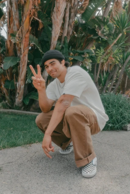 Young man squats outdoors, flashing a peace sign, wearing a beanie, white t-shirt, brown pants, and checkered sneakers, surrounded by lush plants.