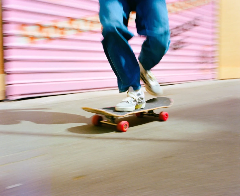 Skateboarder in motion on concrete street, captured with motion blur and red wheels, set against a pink wall with blurred graffiti.