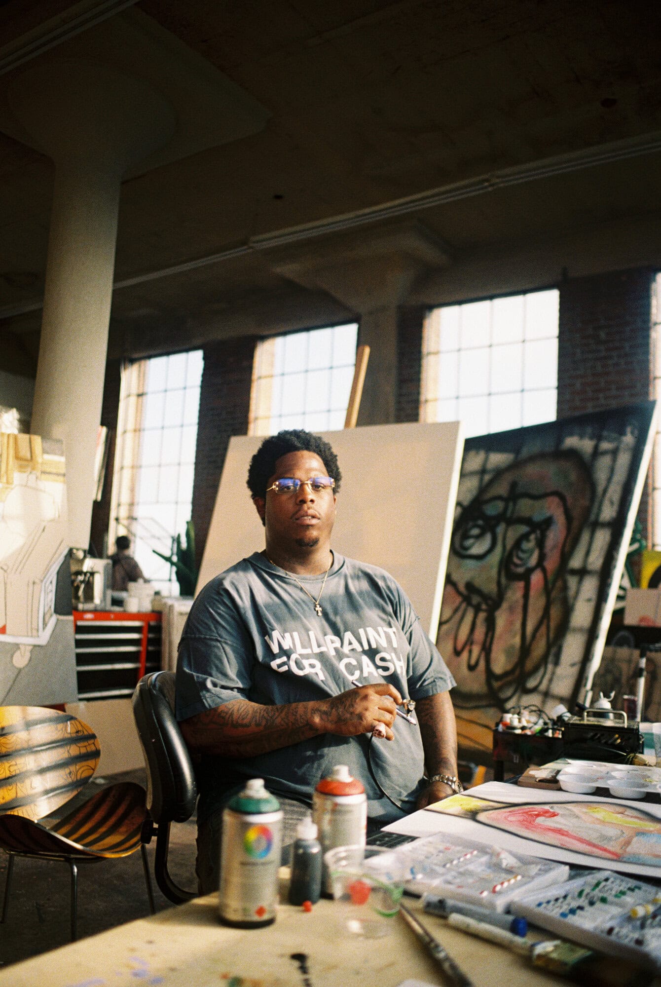 Indoor shot of a man sitting at a desk in front of art canvases in a studio. He is wearing glasses and a t-shirt.
