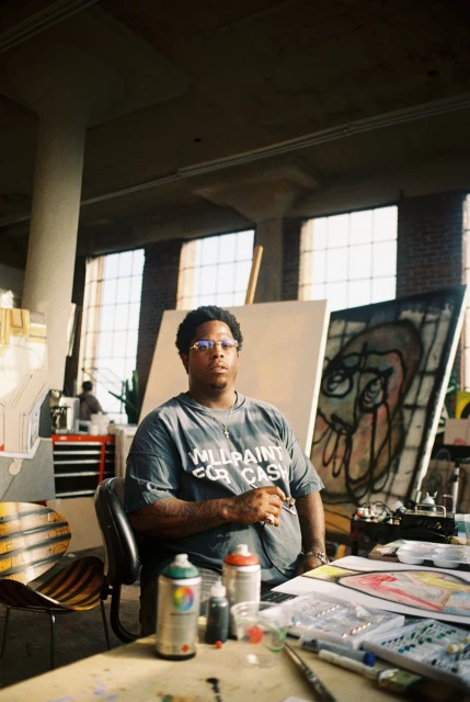 Indoor shot of a man sitting at a desk in front of art canvases in a studio. He is wearing glasses and a t-shirt.