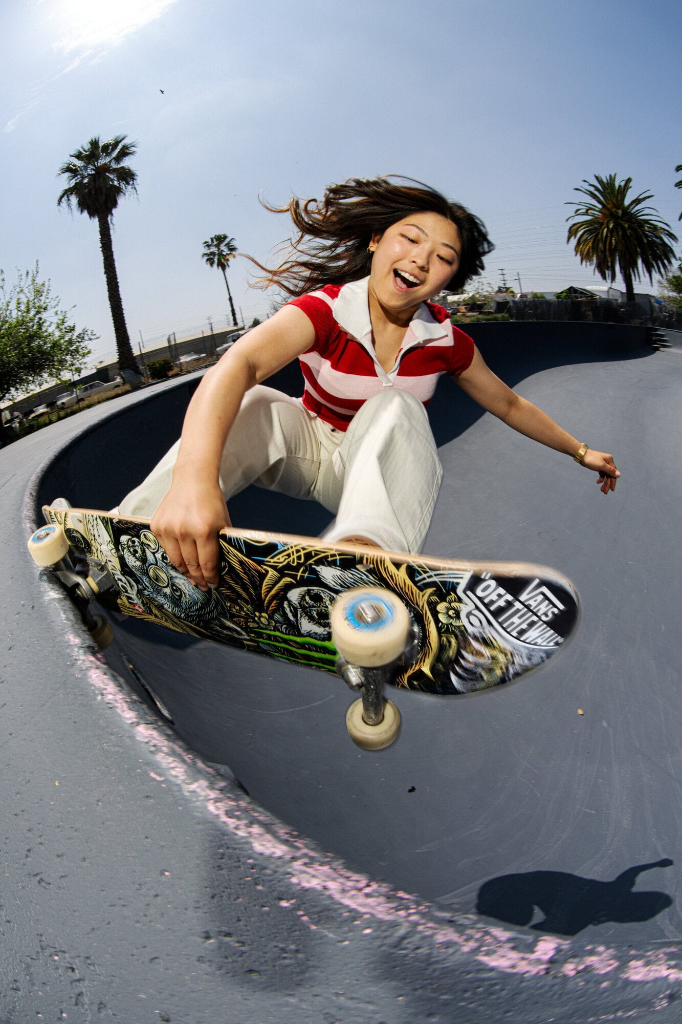 A young woman with long brown hair rides a skateboard on the side of a gray skatepark ramp on a sunny day outside among palm trees.