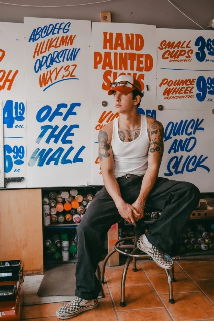 Tattooed man with cap sits in front of hand-painted sign display with typography examples, next to cans of spray paint and checkered shoes.